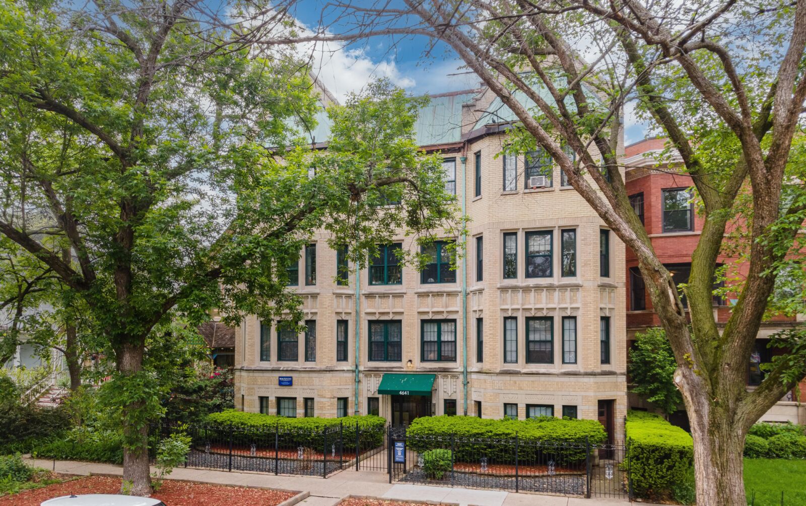 A beige brick apartment building framed by mature trees, with tall bay windows and decorative sto...