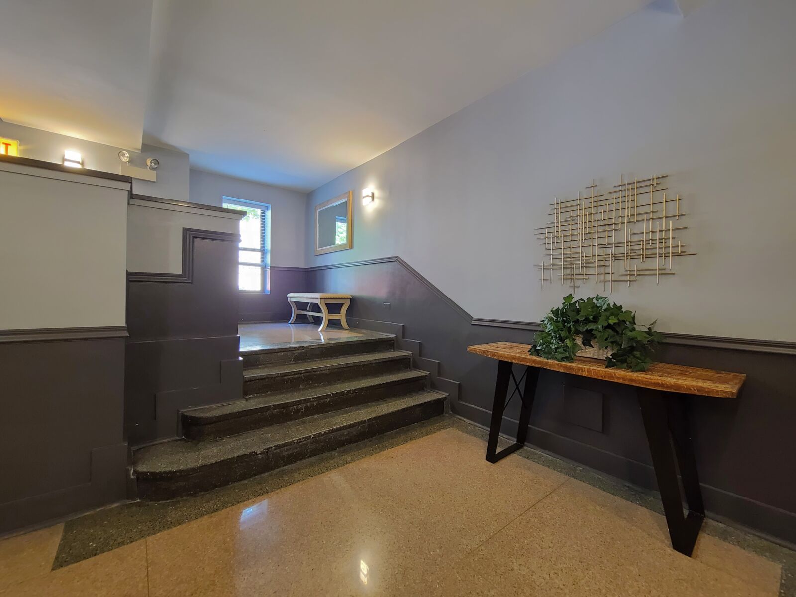 Lobby stairwell with rustic table, potted greenery, and bench seating near bright window.
