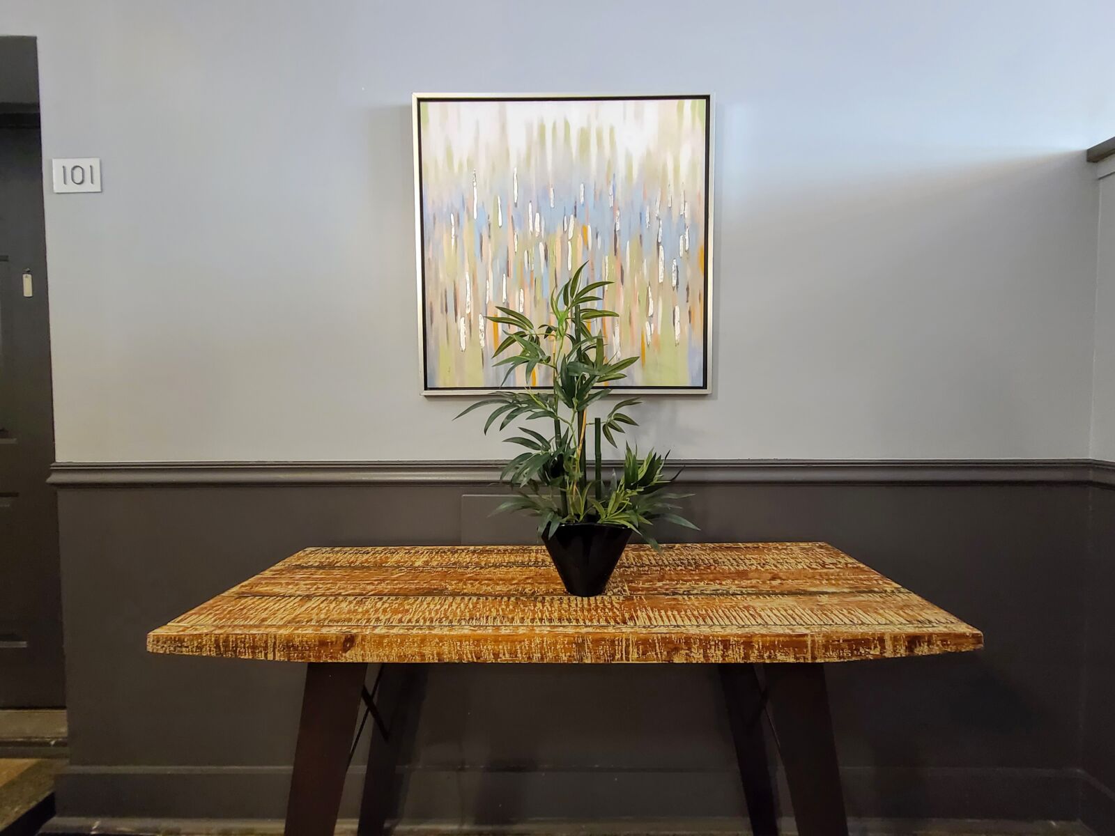 Lobby corner with rustic table, leafy plant in black pot, and colorful abstract wall art.