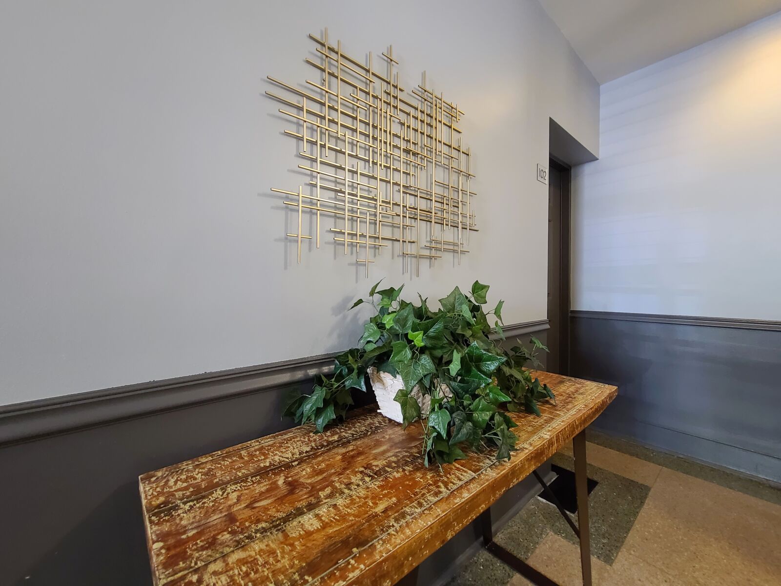Lobby detail with ivy planter on distressed wood table and geometric gold wall sculpture above.