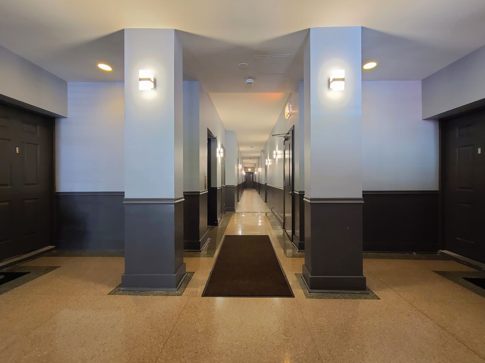 Wide apartment hallway with soft sconces, glossy floors, and dark-trimmed doors lining both sides.