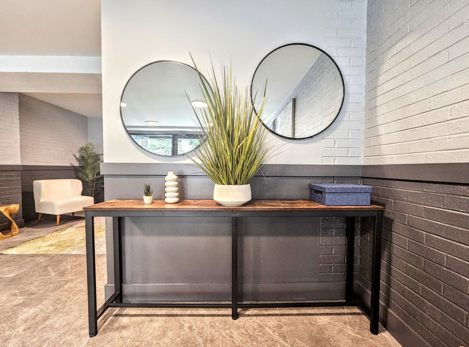Lobby vignette with slim wood console, twin round mirrors, and tall greenery against a two-tone b...