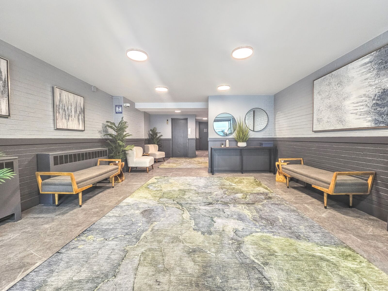 Lobby with gray tile floors, wood-frame benches, and large abstract rug under recessed ceiling li...