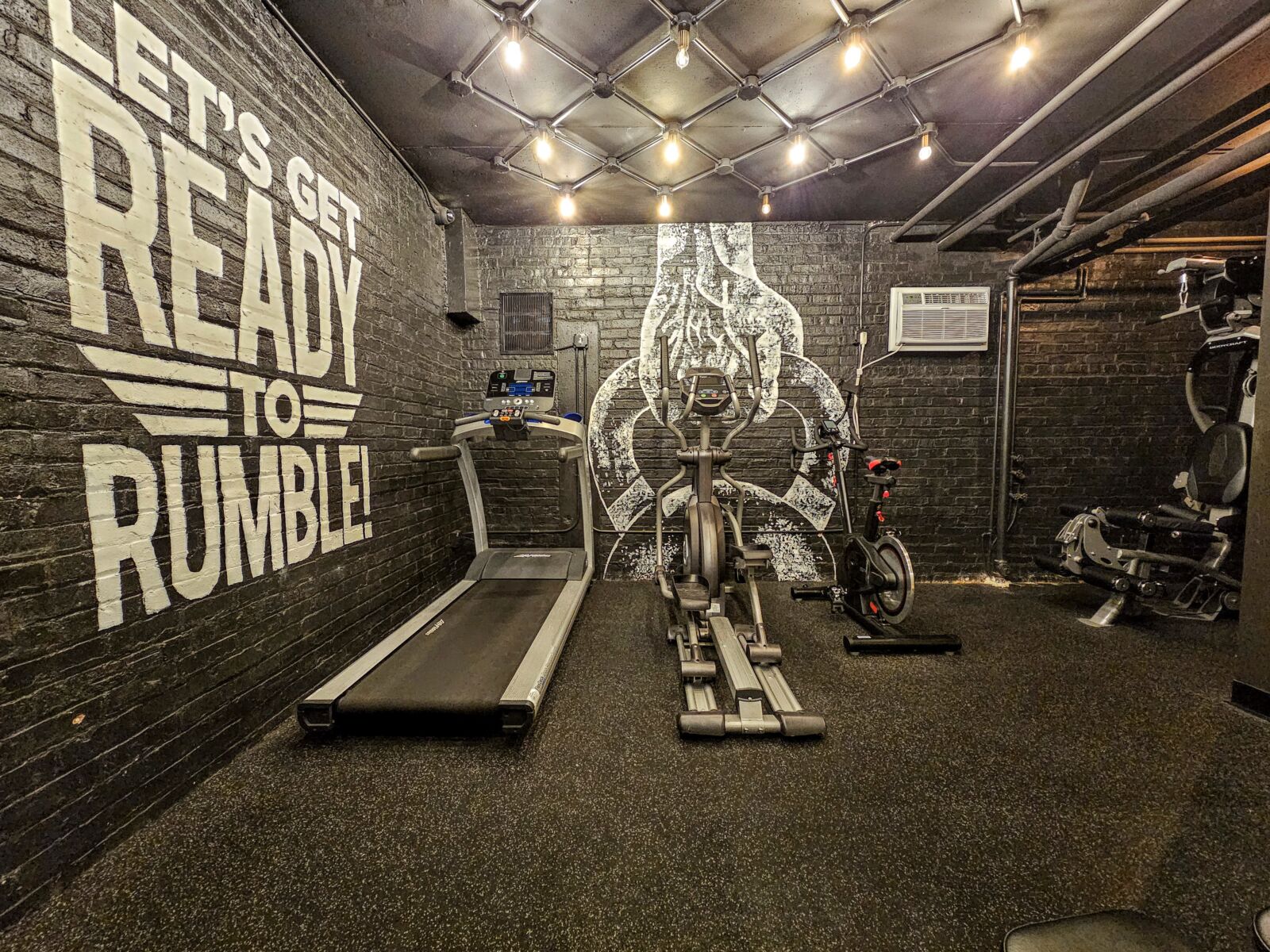 Compact fitness room with a treadmill, elliptical, and stationary bike in front of a black brick ...