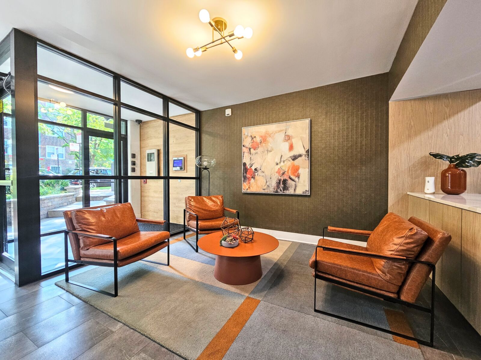 Lobby view with glass walls, abstract art, and modern chandelier above cozy seating arrangement.