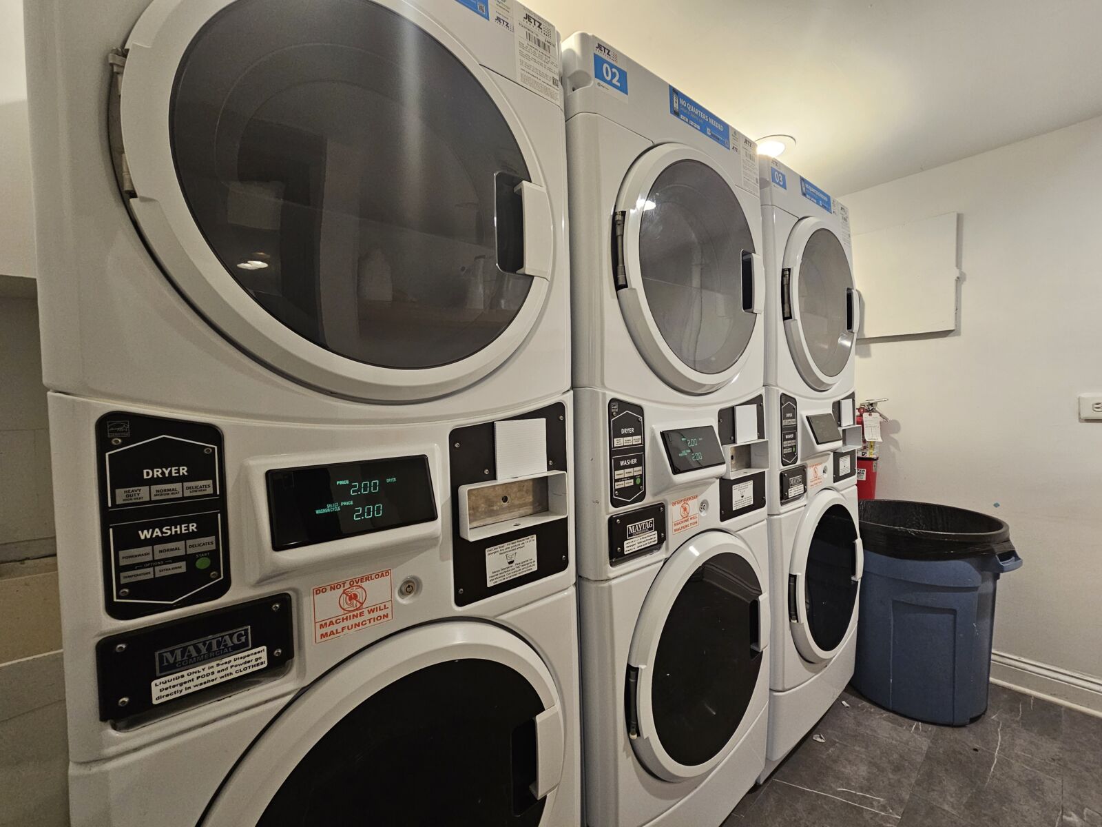 Laundry room with stacked Maytag washers and dryers, digital displays, and nearby trash bin.
