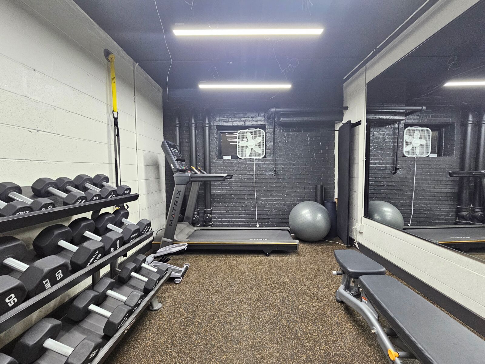 Fitness room with treadmill, dumbbell rack, large mirror wall, and exercise ball on rubber flooring.