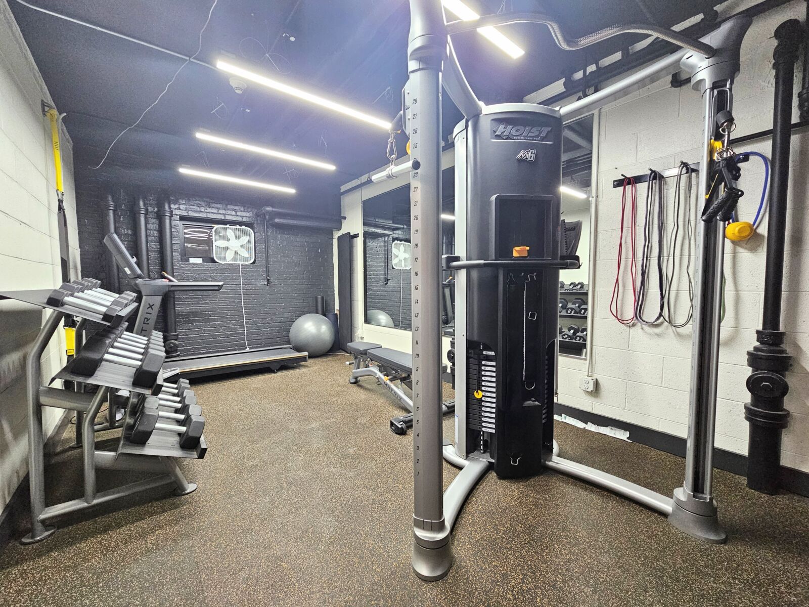 Fitness room with cable machine, resistance bands, dumbbells, and mirrored wall under bright ceil...
