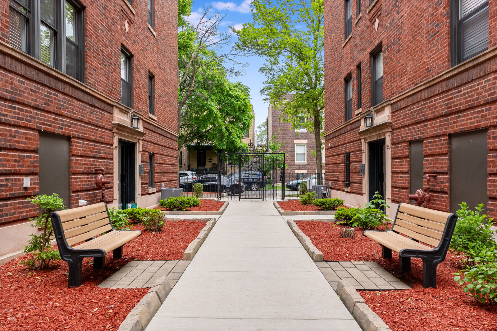 Red brick apartment buildings with gated entrance and landscaped courtyard featuring benches, mul...