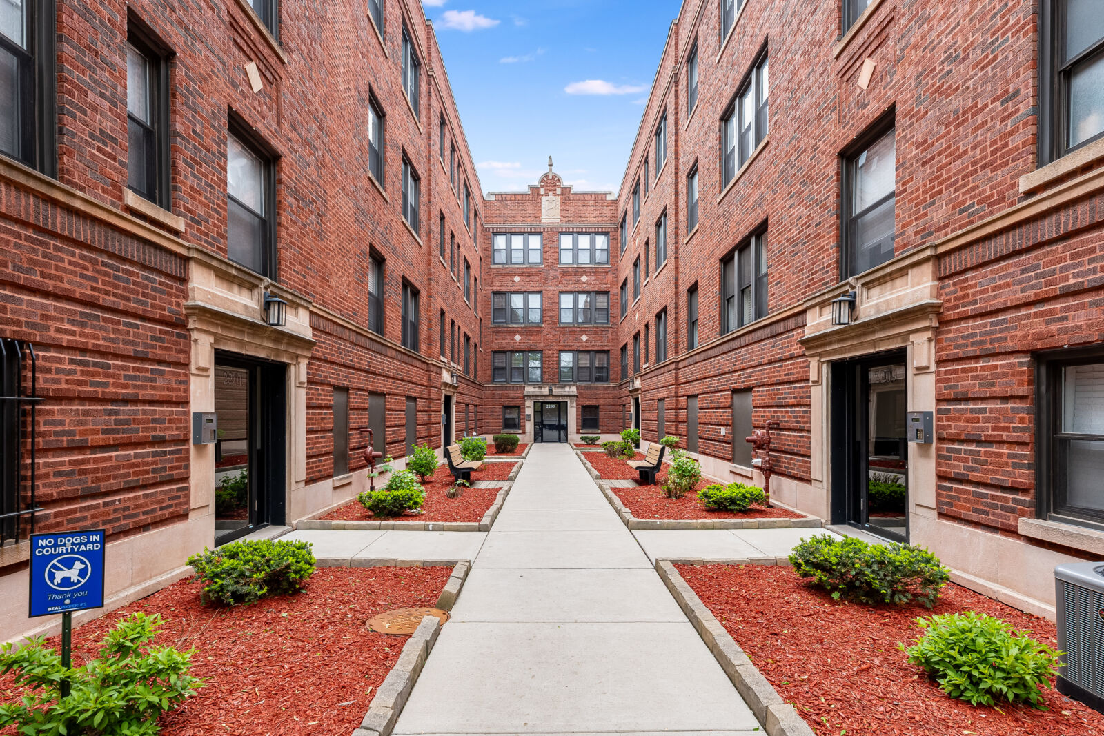 Symmetrical courtyard between two red brick apartment buildings, with benches, trimmed shrubs, an...