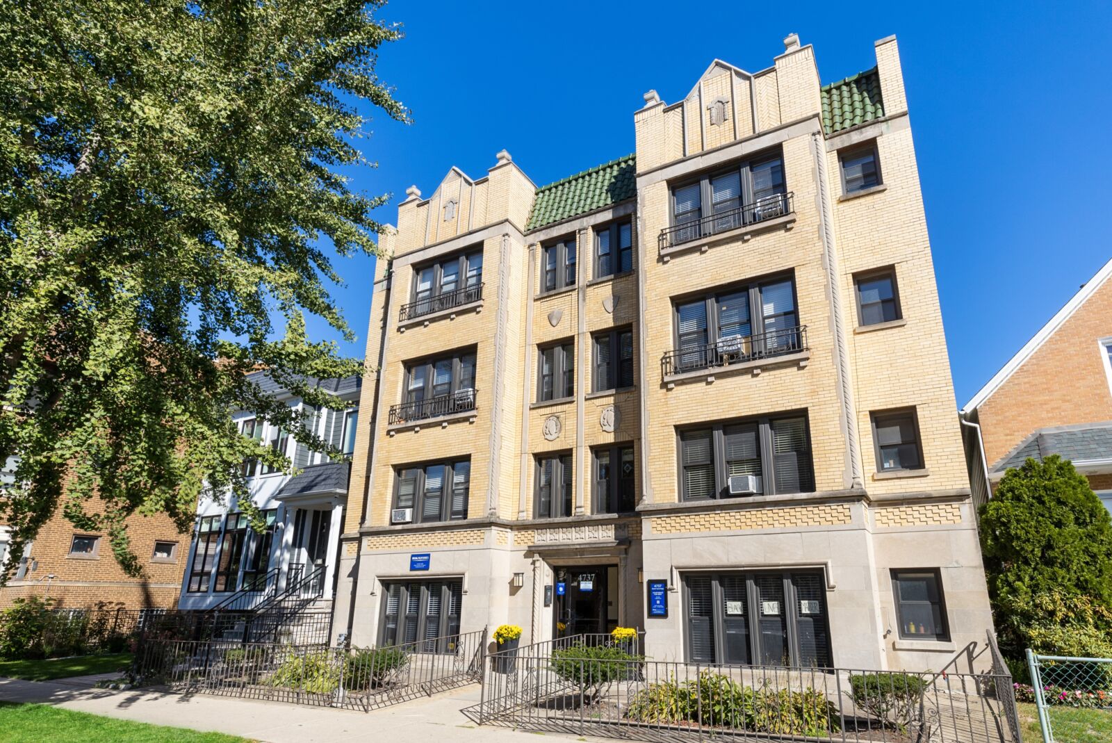 Four-story tan brick building with green tile accents, decorative stonework, and gated entry garden.