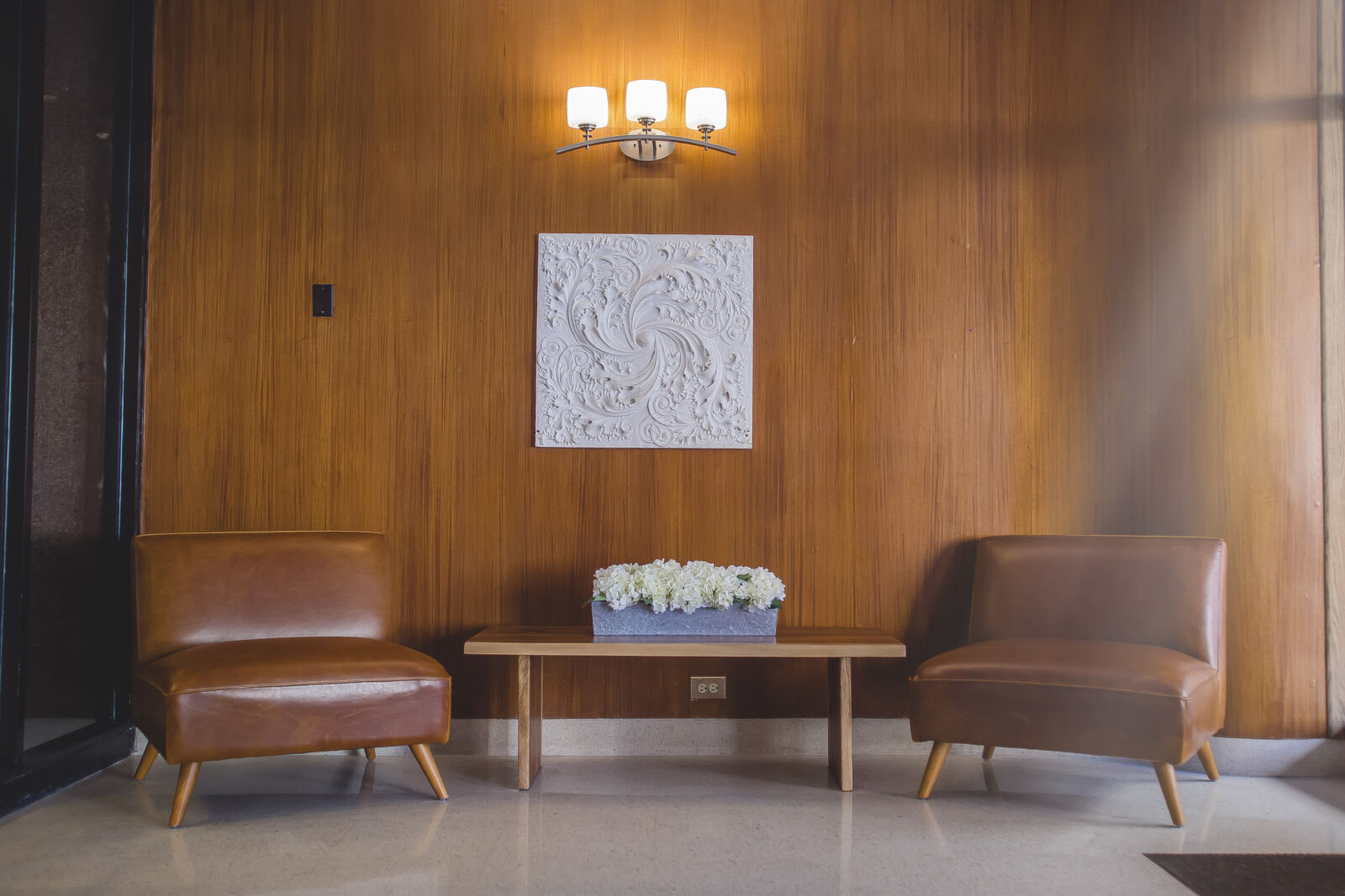 Lobby with mid-century modern chairs, wood-paneled wall, and wall sconce lighting above floral de...