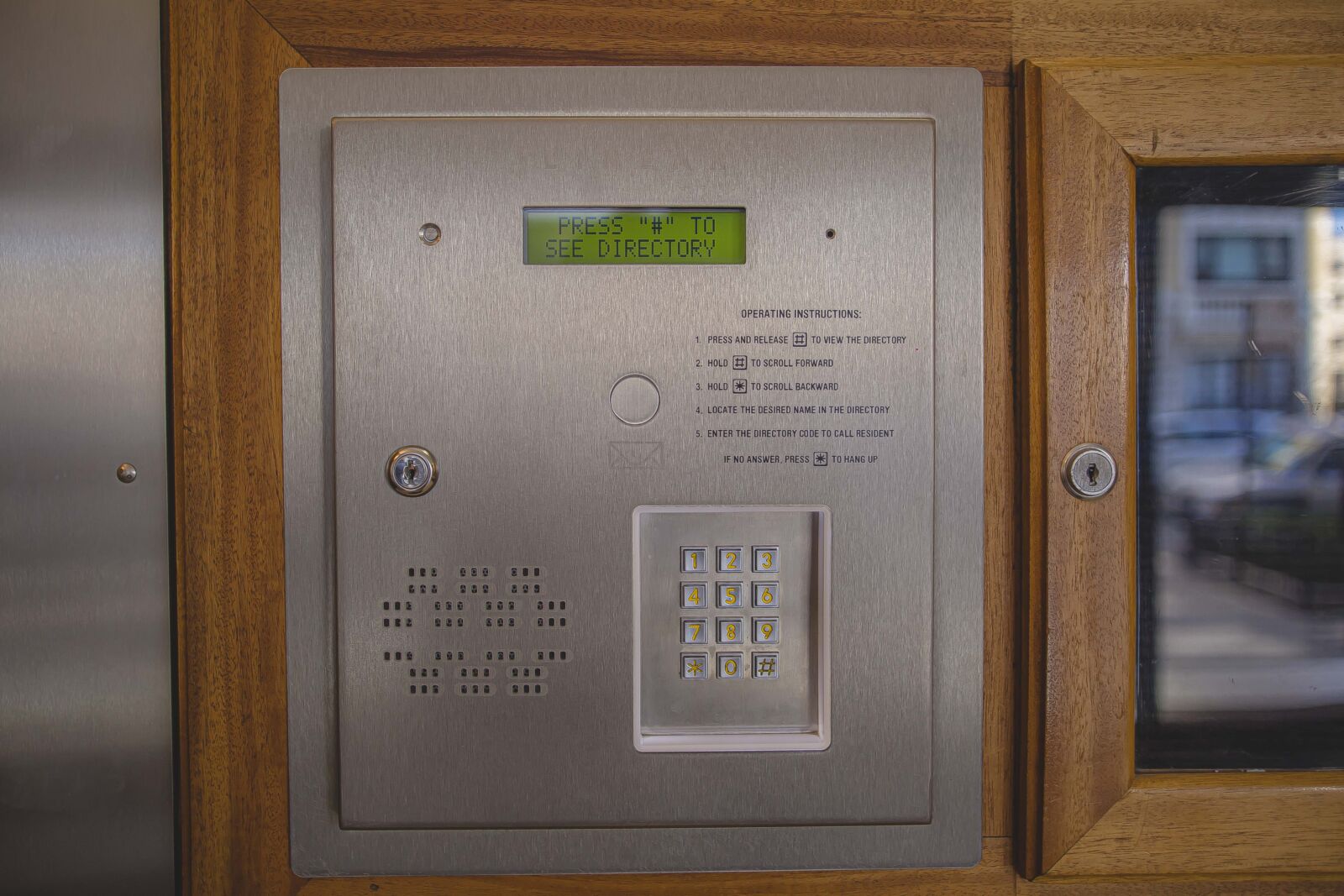 Building entry intercom with keypad and digital directory screen set in a wood-framed panel.