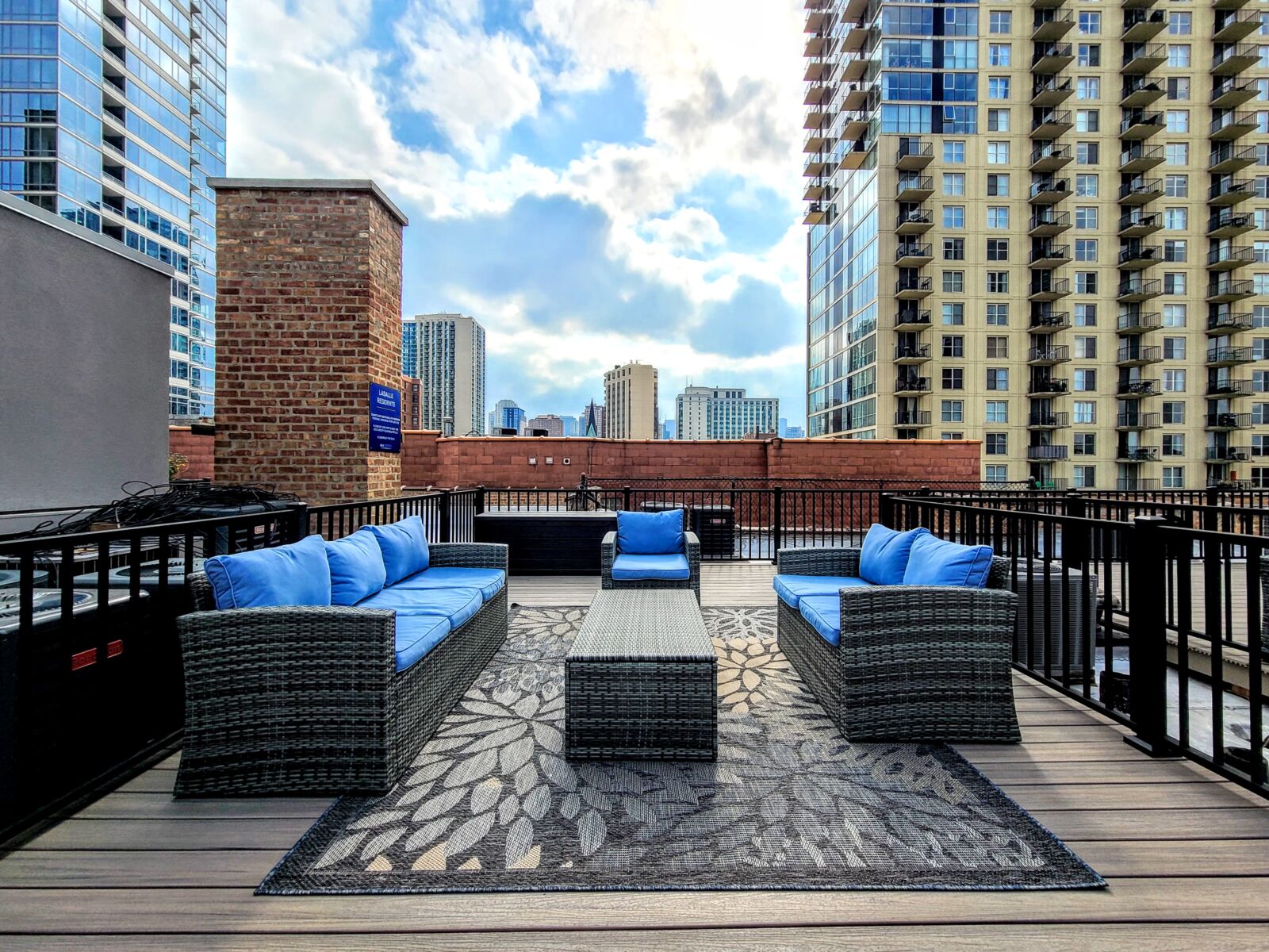 Rooftop lounge with wicker seating, blue cushions, and skyline views framed by high-rise buildings.